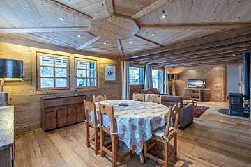 Inviting rustic chalet interior with extensive wooden paneling, an oval dining table, a lounge area with TV, and a classic wood stove.