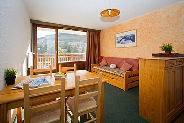 A cozy, brightly lit living-dining room with wooden furniture, orange walls, and a green carpet. It features a balcony offering a scenic mountain view.