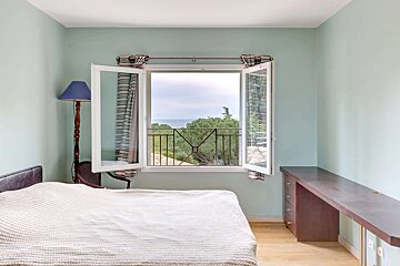 Light blue bedroom with an open window revealing a sea and tree view. A bed, desk, and floor lamp furnish the space.
