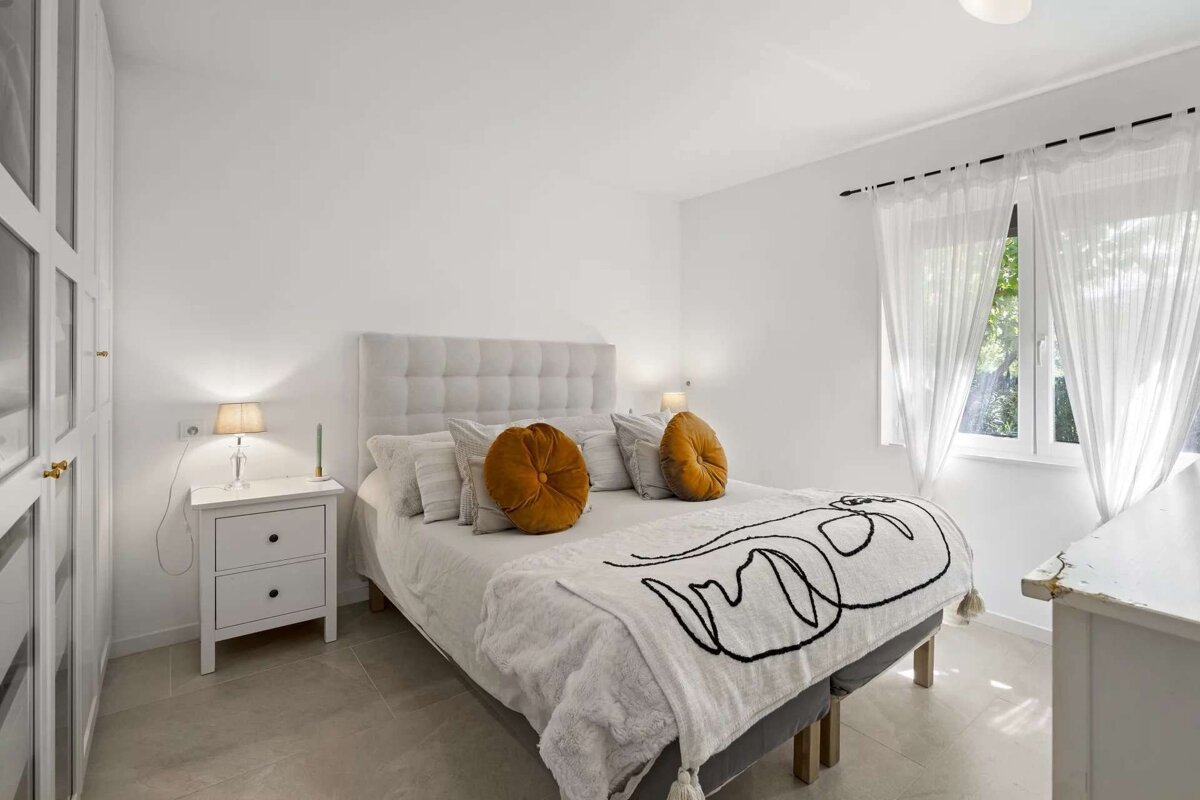 A bright, minimalist white bedroom features a bed with orange accent pillows, a graphic throw blanket, a nightstand, and a sunlit window with sheer curtains.