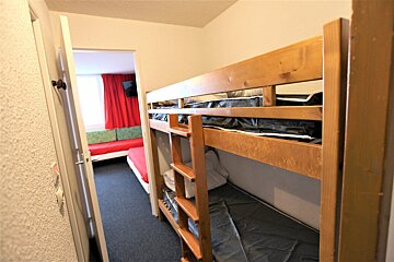 A room with wooden bunk beds in the foreground. Through an open doorway, another room with red curtains and red/green sofa beds is visible.