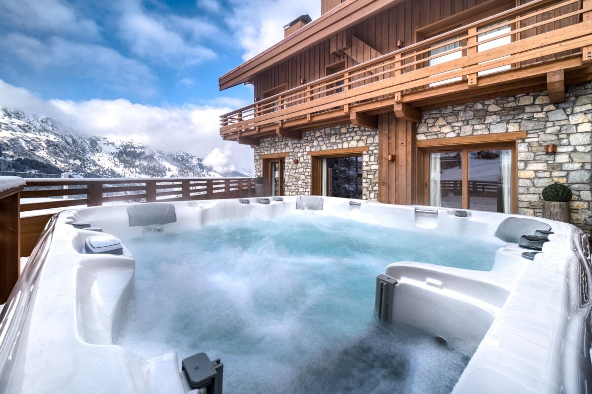 A steaming outdoor hot tub on a snowy mountain terrace, backed by a rustic chalet and snow-covered peaks under a partly cloudy sky.