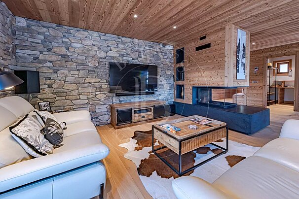 Rustic-modern living room with a stone wall, wood paneling, white leather sofas, a sleek fireplace, and a cowhide rug on a wooden floor.