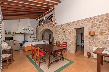Spacious rustic living/dining room with stone walls, exposed wooden beams, a large dining table, and a cozy lounge area with a fireplace. A mezzanine overlooks the space.