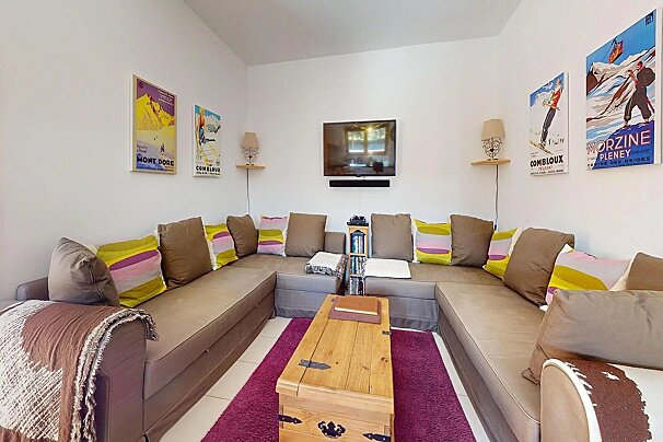 Cozy living room featuring a large U-shaped sofa with colorful pillows, ski-themed wall art, a TV, and a wooden trunk coffee table on a purple rug.