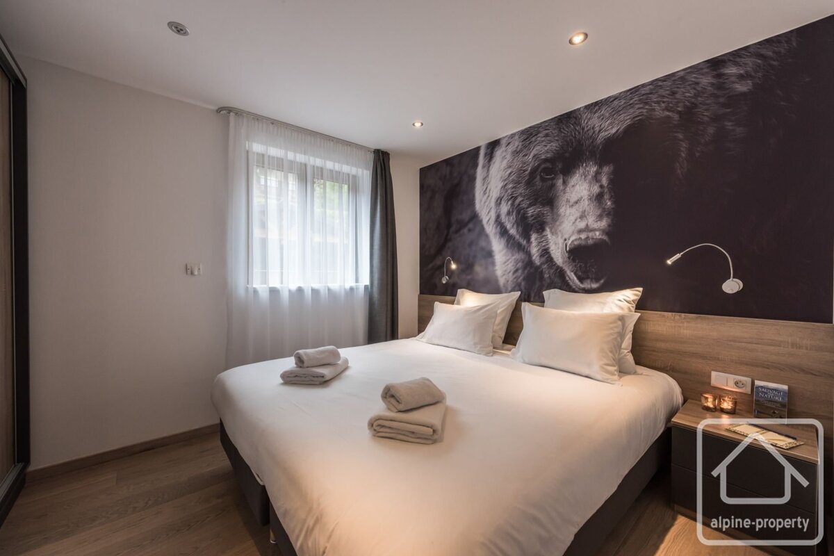A modern bedroom features a large bed with white linens, a window with sheer curtains, and a striking dark mural of a bear's face behind the headboard.