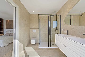Modern beige-tiled bathroom with a shower, floating toilet, white vanity, and abstract mirror, connected to a bedroom via a sliding door.