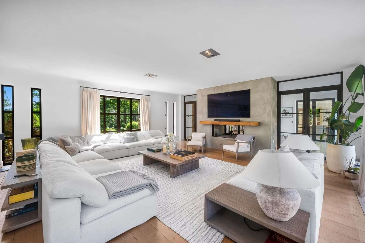 A spacious, modern living room featuring a white sectional, large TV over a fireplace, wooden floors, and lush green views from expansive windows.