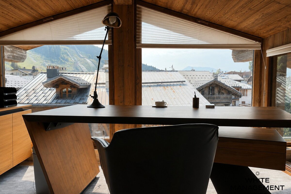 A cozy chalet home office with a wooden desk facing large windows that offer views of alpine rooftops and mountains.