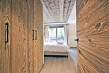 Rustic chalet bedroom with wood paneling, a double bed with white linens, and a large wardrobe, viewed through an open wooden door.