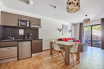 A modern open-plan apartment featuring a compact kitchen, dining table with white chairs, a red sofa, wood floors, and balcony access.