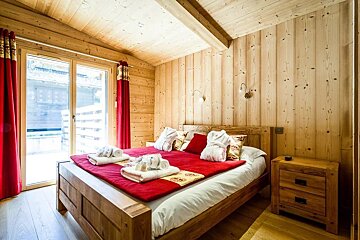 Cozy wooden chalet bedroom featuring a bed with red and white linens, matching curtains, and wood-paneled walls.