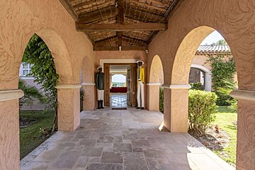 A peach-toned arched outdoor hallway with a stone floor and beamed ceiling. Two statues stand before an open door, with greenery and gardens visible beyond.
