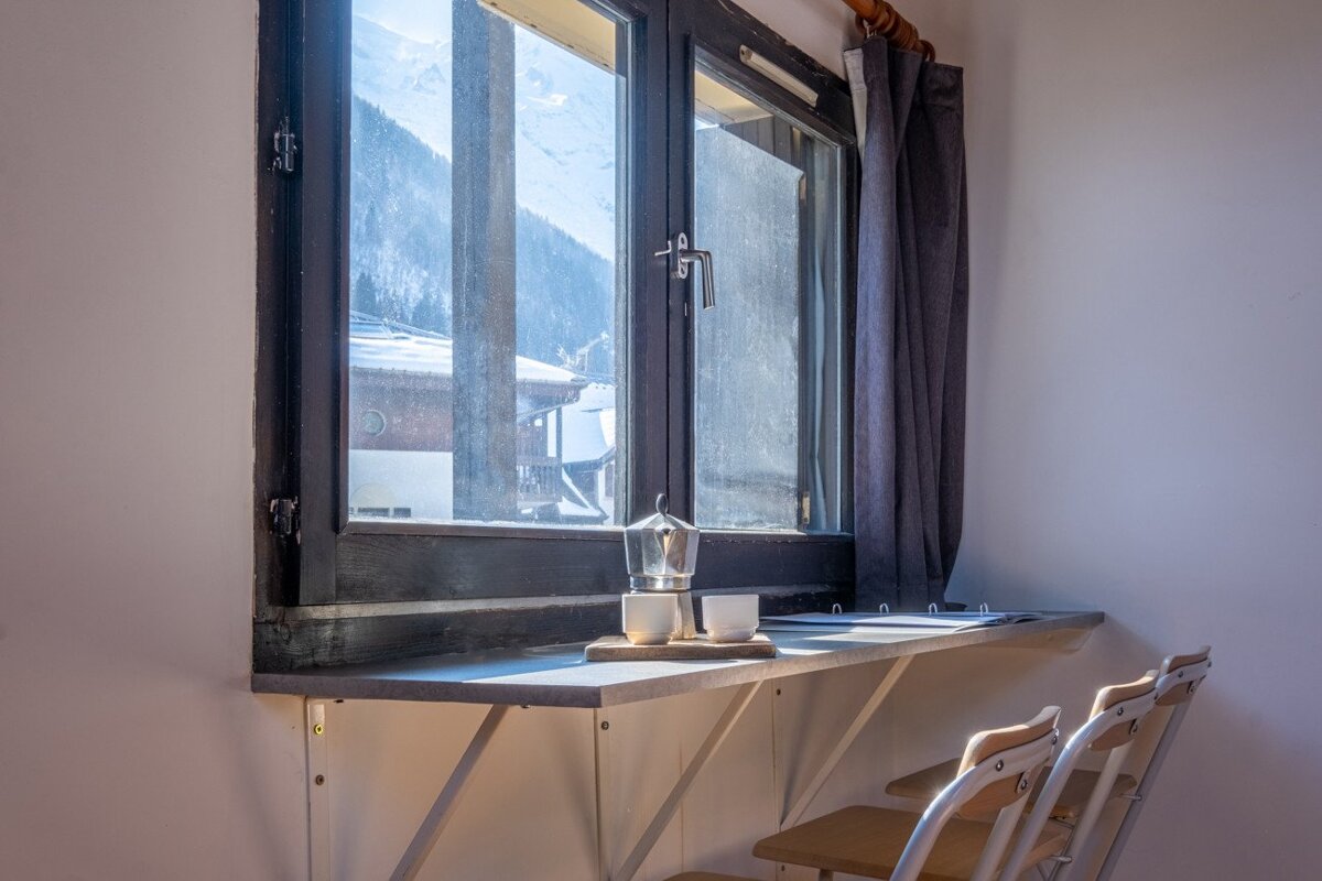A window overlooking a snowy mountain village, with a bar holding a moka pot and two cups, and chairs below.