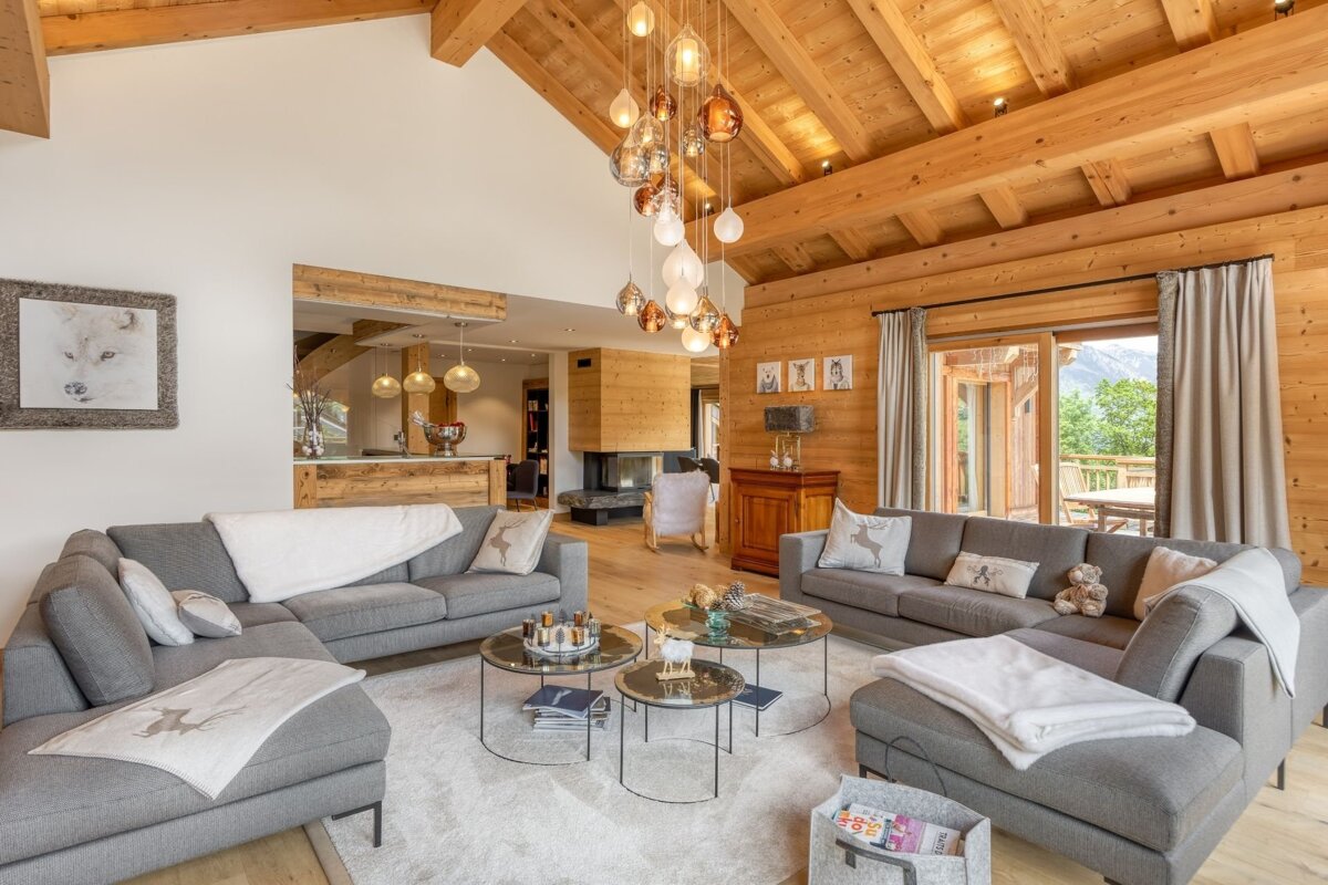 Modern chalet living room with wood-paneled walls/ceiling, grey sofas, a unique cascading chandelier, and mountain views.