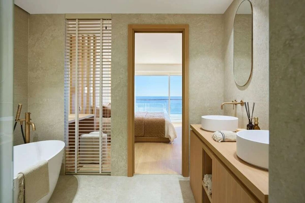 A modern bathroom with a freestanding tub, dual sinks, and a slatted partition connecting to a bedroom with an ocean view.