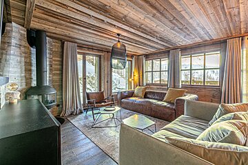A cozy, wood-paneled chalet living room with a modern fireplace, comfy sofas, and large windows showcasing a snowy winter scene.