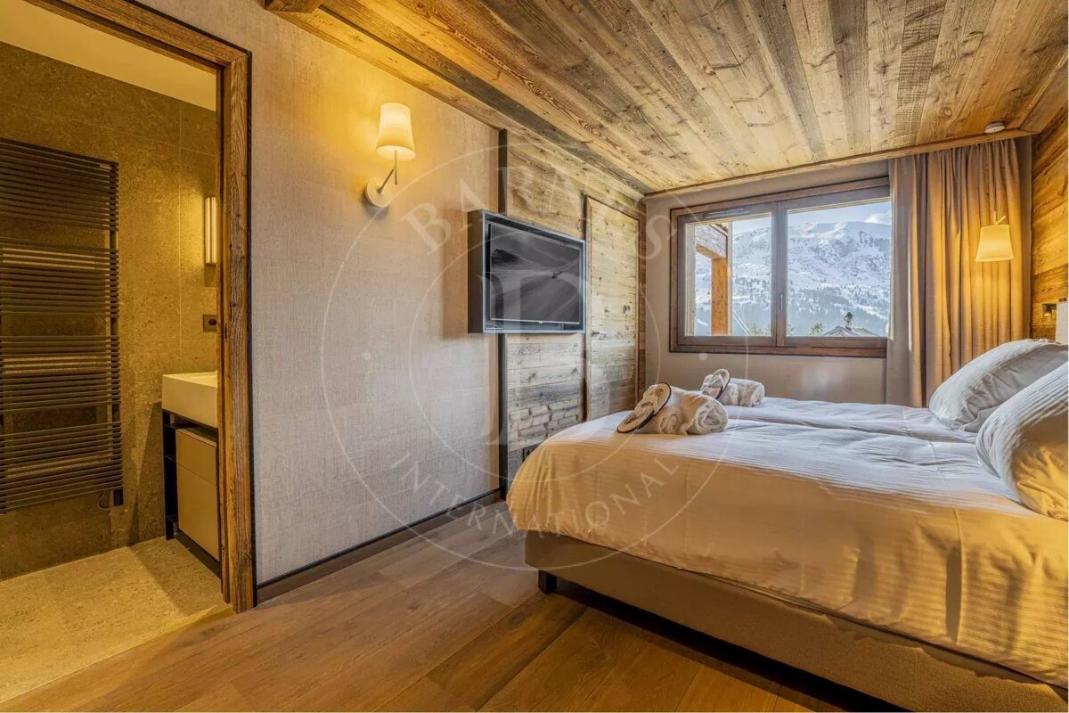 A rustic chalet bedroom featuring a cozy bed, wooden ceiling, mountain view from the window, TV, and an open doorway to a modern bathroom.