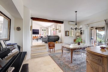 A bright, multi-level home with a dining area, piano, and a sunken living room visible through a stepped opening. Outdoor view through a doorway.
