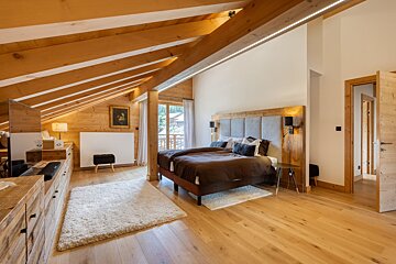A cozy, light-filled attic bedroom with exposed wooden beams, twin beds, and a balcony view. Features warm wood finishes and modern lighting.