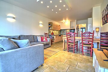 A bright, open-plan living space features a grey L-shaped sofa, a dining area with a wooden table and red chairs, and a compact kitchenette, all on a tiled floor.