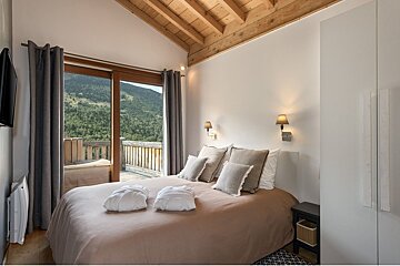 A cozy bedroom with a mountain view balcony, wooden ceiling, and modern decor, featuring a bed with robes, sconces, and a white wardrobe.