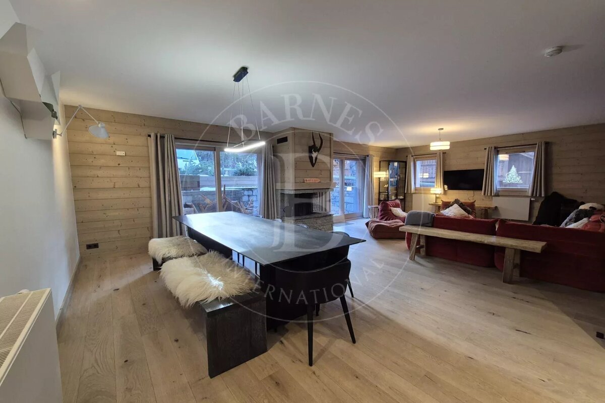 A cozy modern chalet interior featuring a dining table with sheepskin benches and a living area with a fireplace and red sofas.