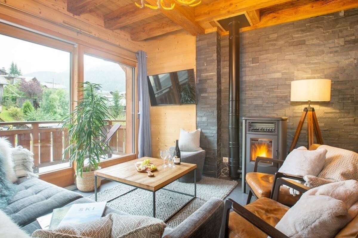 Cozy chalet living room with rustic wooden beams, a stone fireplace, leather armchairs, and large windows showing a scenic outdoor view.