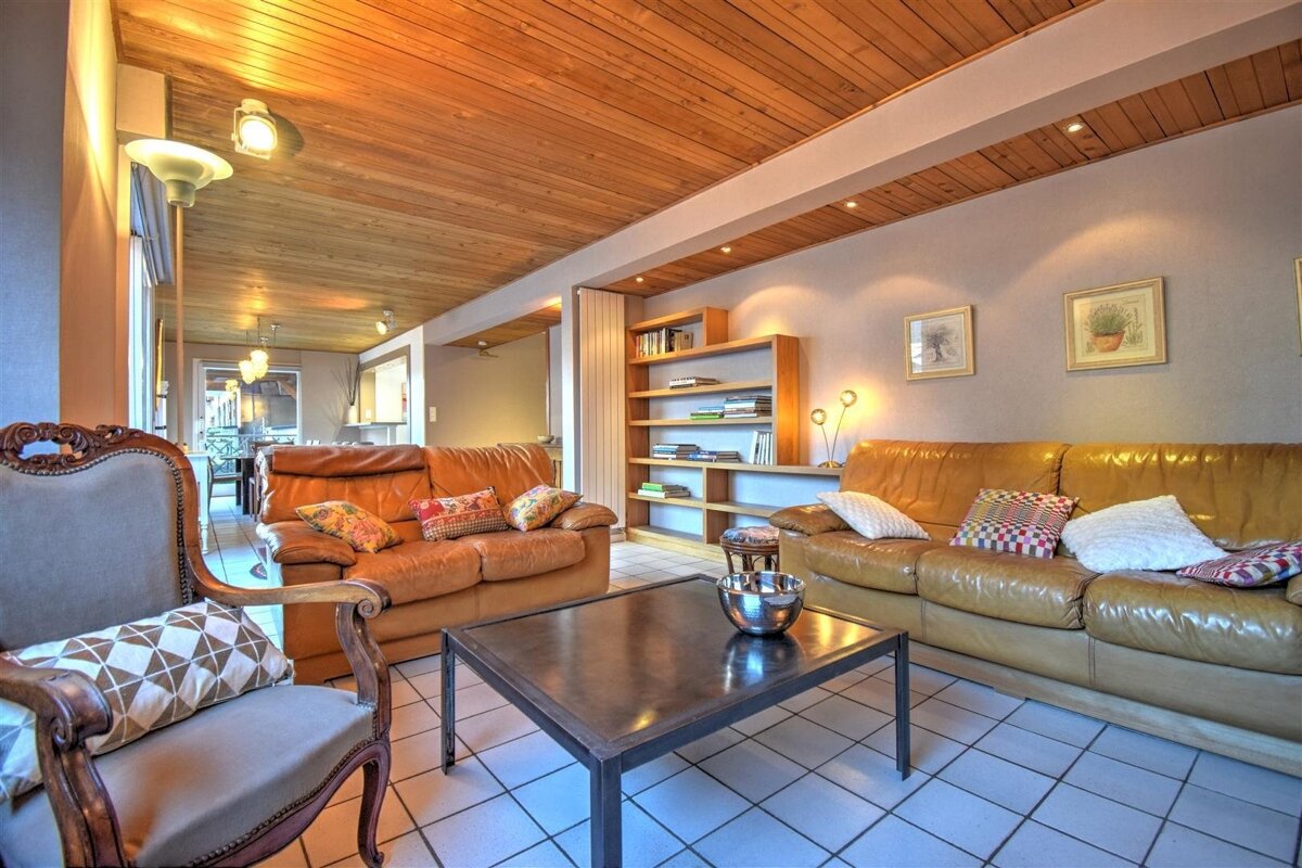 A warm living room features a wooden plank ceiling, tan leather sofas, a dark coffee table, tiled floor, and a bookshelf with framed artwork.