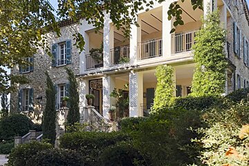 An elegant stone villa with blue shutters and a white pillared terrace, surrounded by lush green gardens.