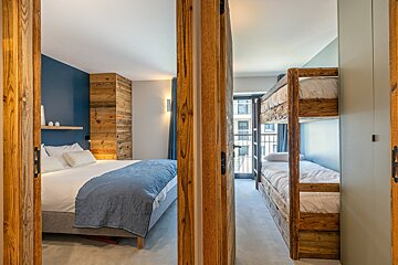 Two cozy bedrooms seen through wooden doorways: one with a double bed and blue accents, and another with rustic wooden bunk beds.