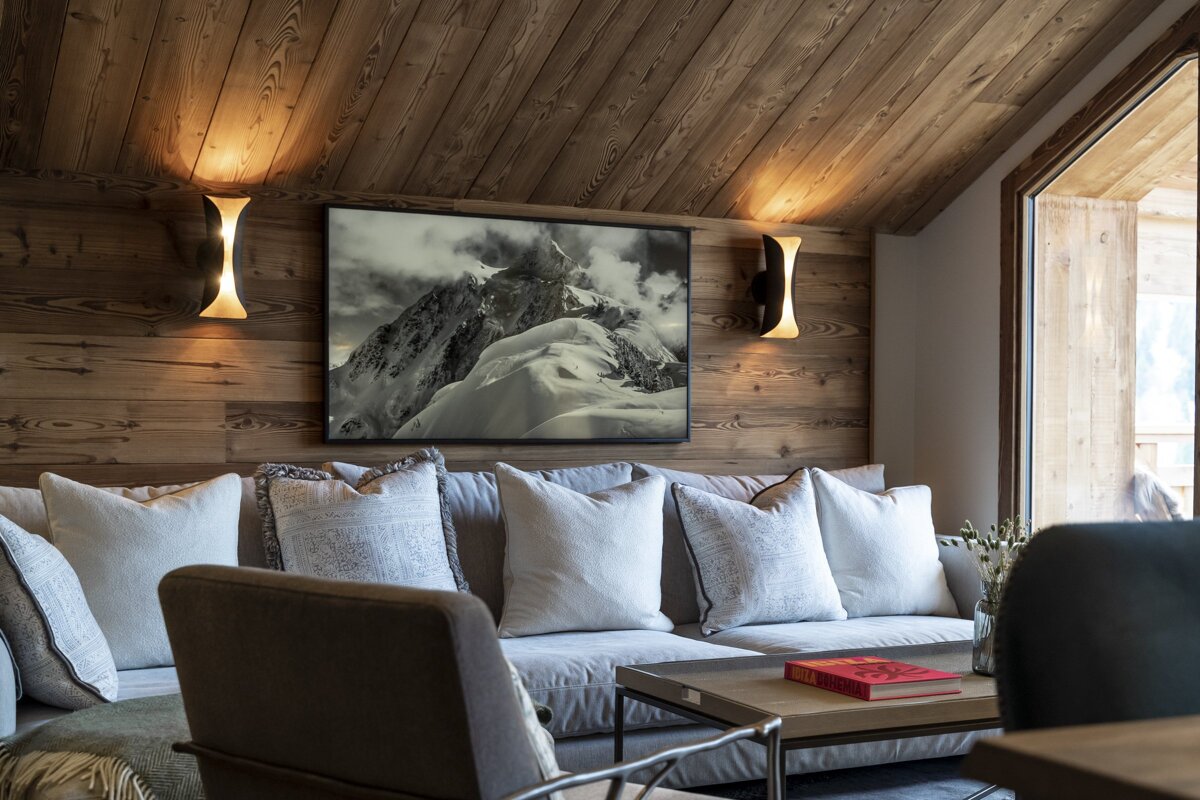 Cozy rustic cabin living room with wood paneled walls, warm sconces, a plush sofa with pillows, and a framed mountain print.