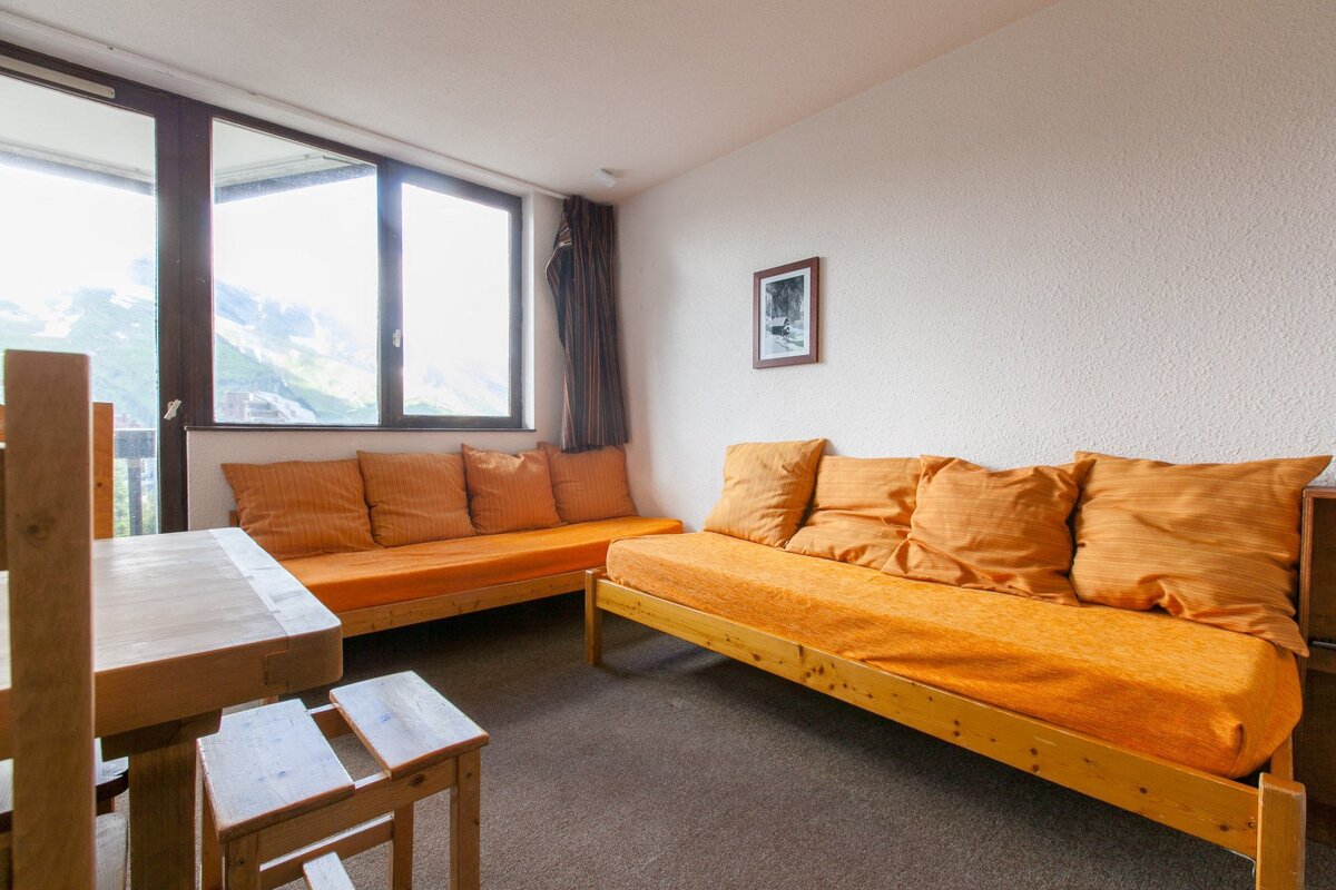 A bright living room with orange daybeds, wooden dining furniture, and large windows showcasing a stunning mountain vista.