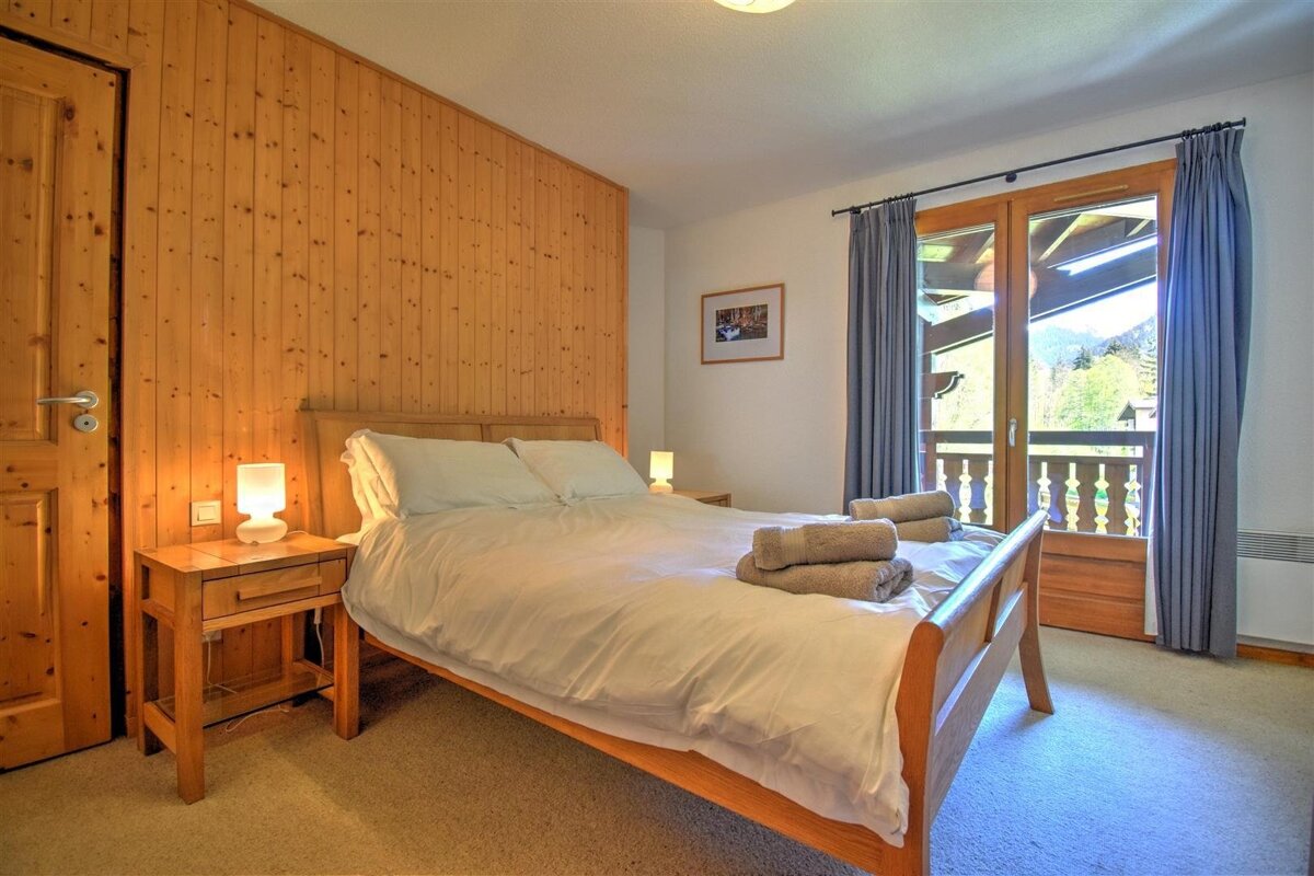 A cozy bedroom with a wooden accent wall, large bed, nightstands, and a balcony door overlooking a scenic mountain landscape.