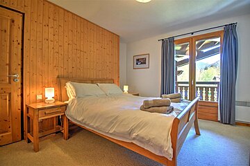 A cozy bedroom with a wooden accent wall, large bed, nightstands, and a balcony door overlooking a scenic mountain landscape.