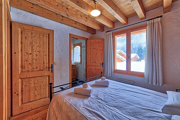 A cozy bedroom featuring exposed wooden beams, white bedding, and a window overlooking a snowy mountain landscape with chalets. Wooden doors are visible.