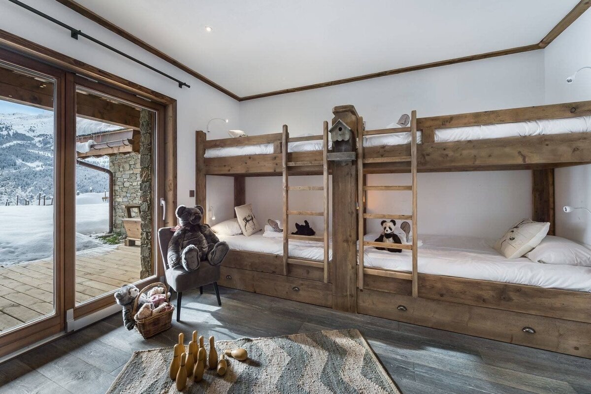 A rustic wooden bunk room with a snowy mountain view from large glass doors, featuring multiple beds, stuffed animals, and a cozy rug.