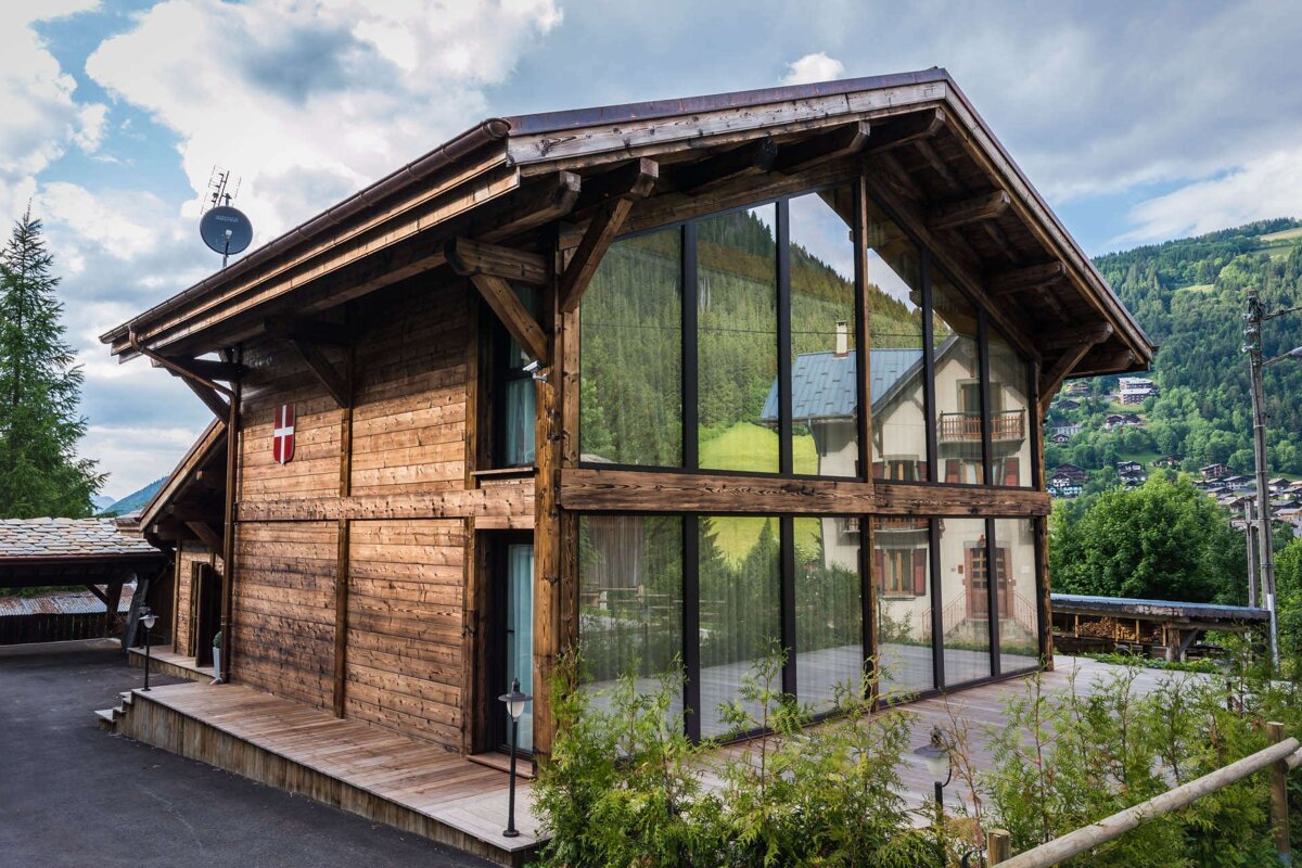 A striking modern wooden chalet with a large glass facade reflecting green mountains and a nearby house, situated in a scenic valley.