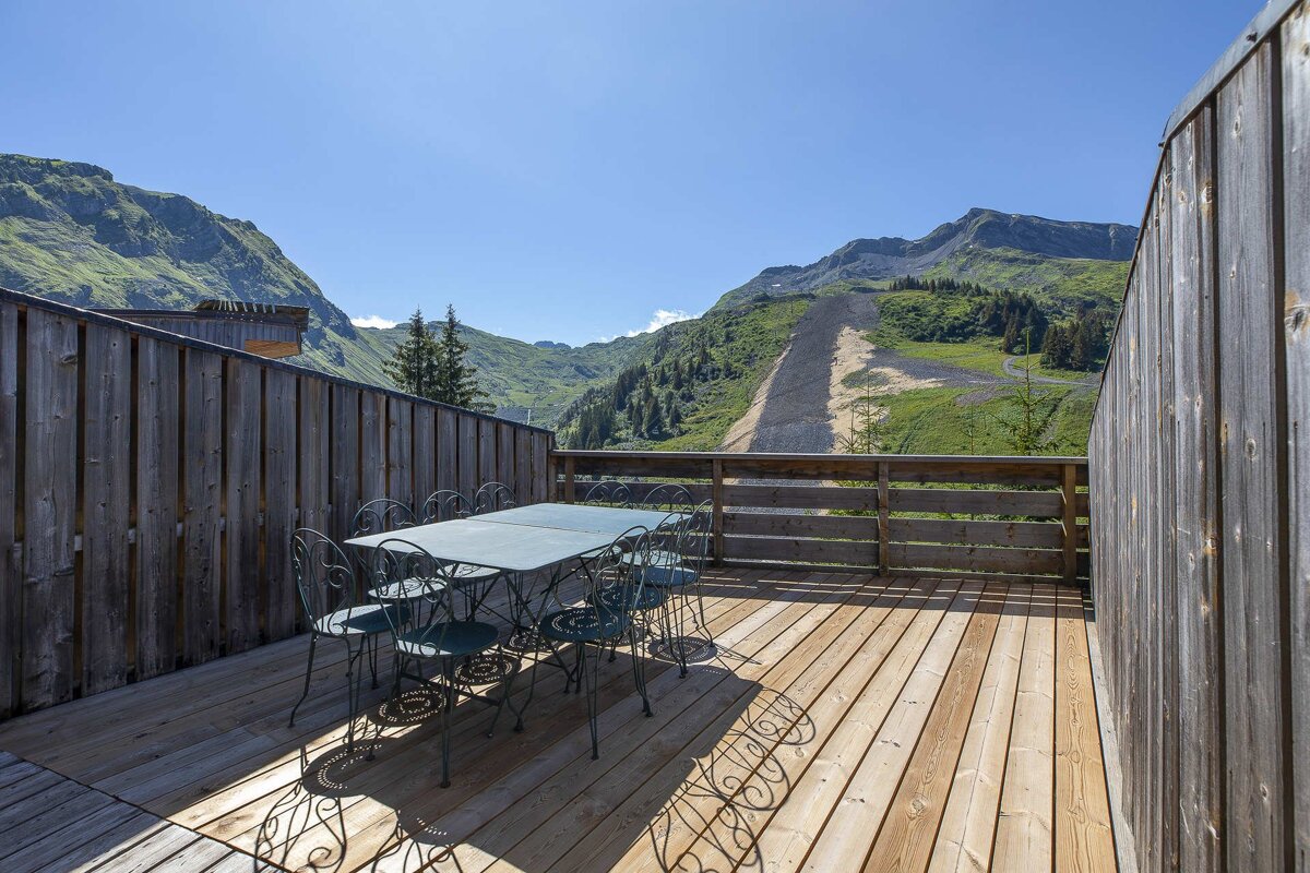 Wooden deck with an outdoor dining set overlooking a stunning green mountain landscape under a clear blue sky.