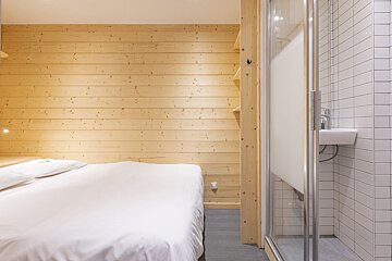 A modern, compact room with a white bed, wooden walls, and a shower enclosure with white tiled walls and a small sink.