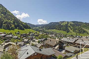 A scenic mountain village nestled among lush green hills and forested peaks, with distant ski slopes visible under a clear blue sky.