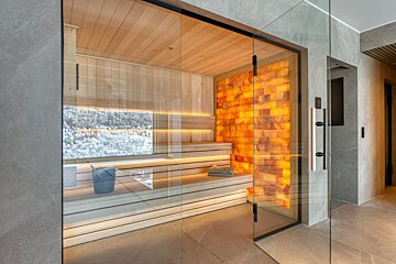 Modern glass sauna with wooden benches, a glowing salt wall, and a snowy forest view.
