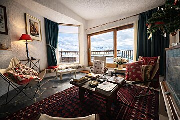 A cozy, rustic living room with large windows overlooking snow-covered mountains, featuring butterfly chairs and patterned rugs.