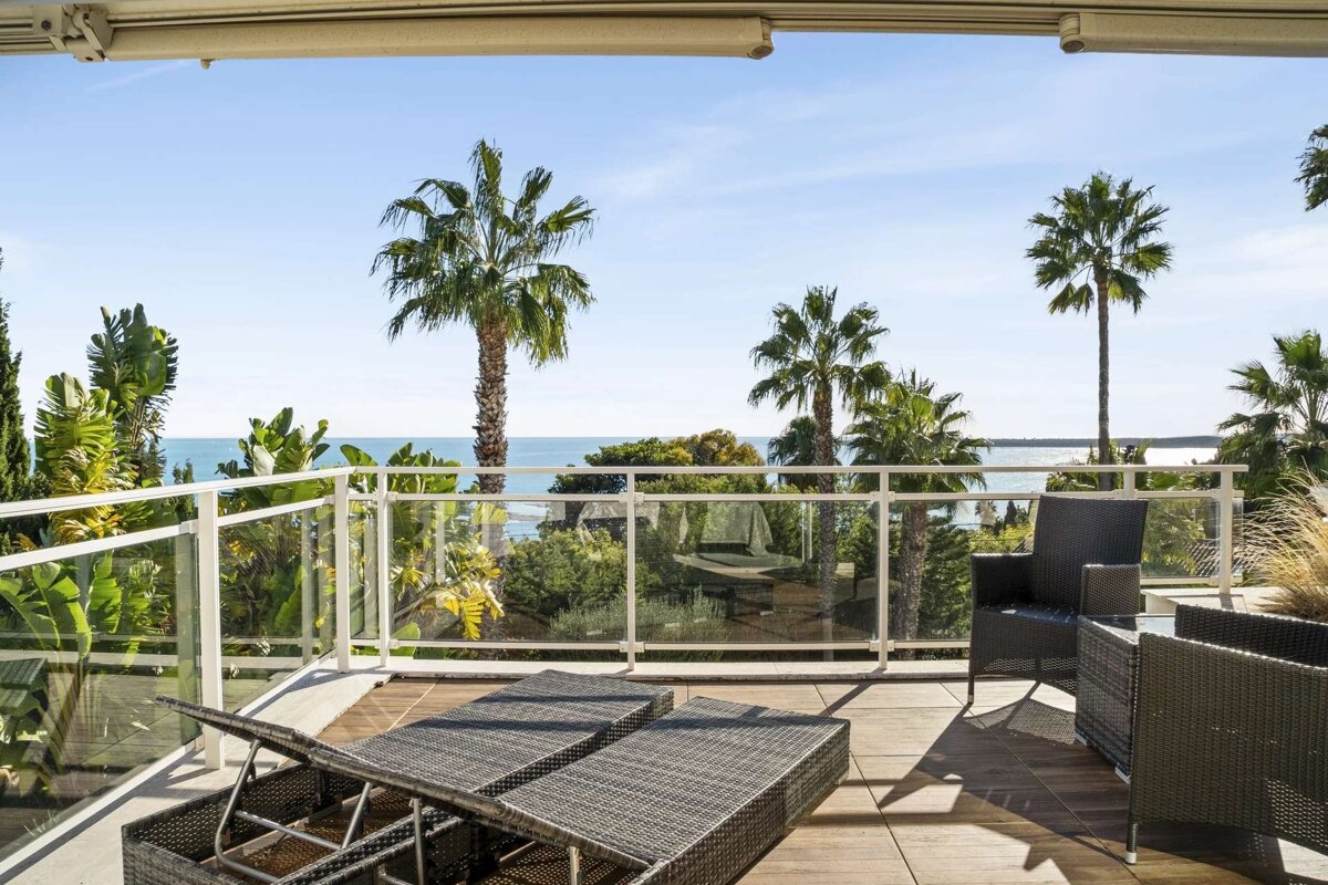 A sunny balcony with wicker lounge chairs and seating offers a beautiful view of the blue ocean, framed by lush palm trees under a clear sky.