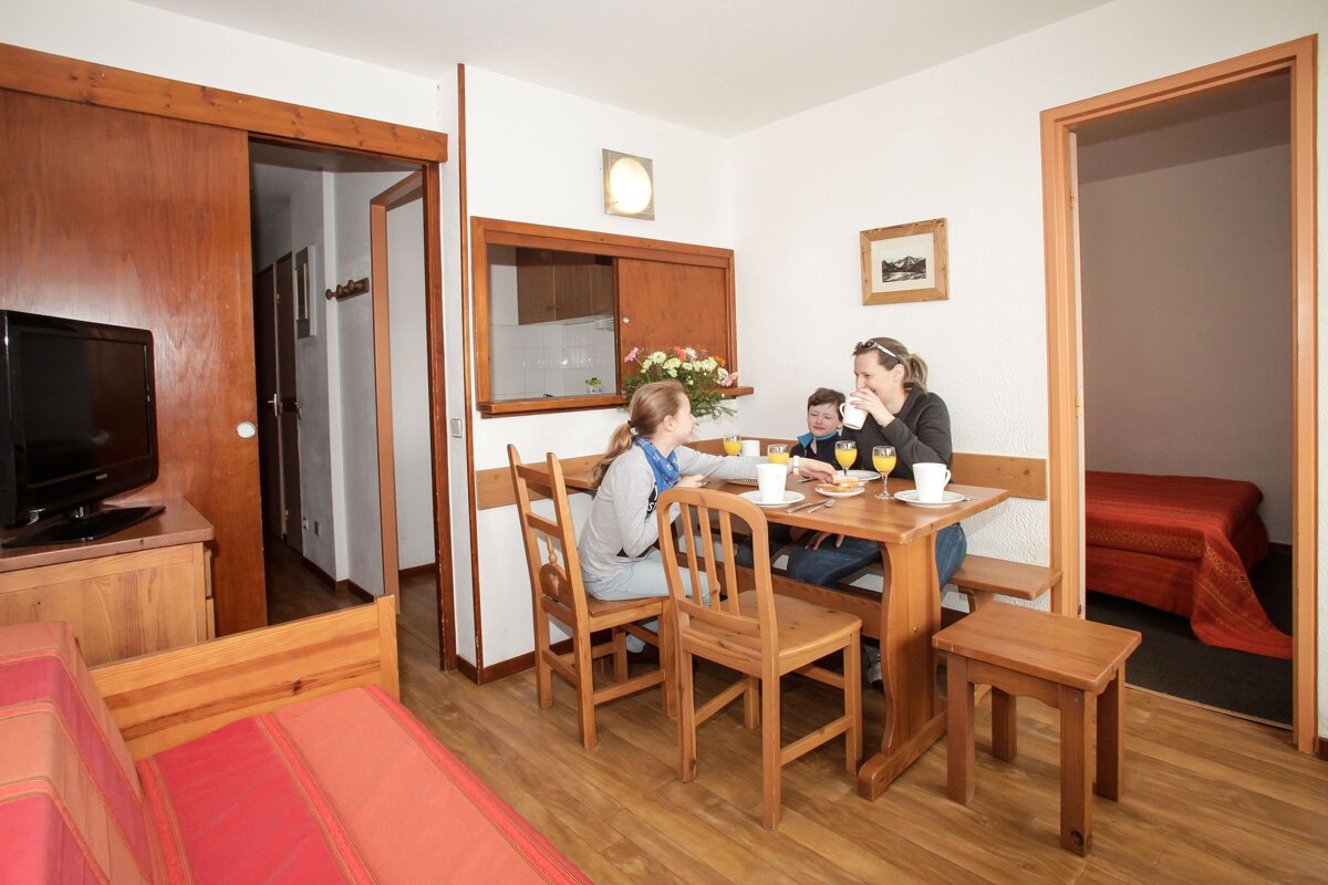 A family of three enjoys a meal at a wooden table in a bright, wood-themed apartment, showing dining, living, and bedroom areas, with a kitchen pass-through.