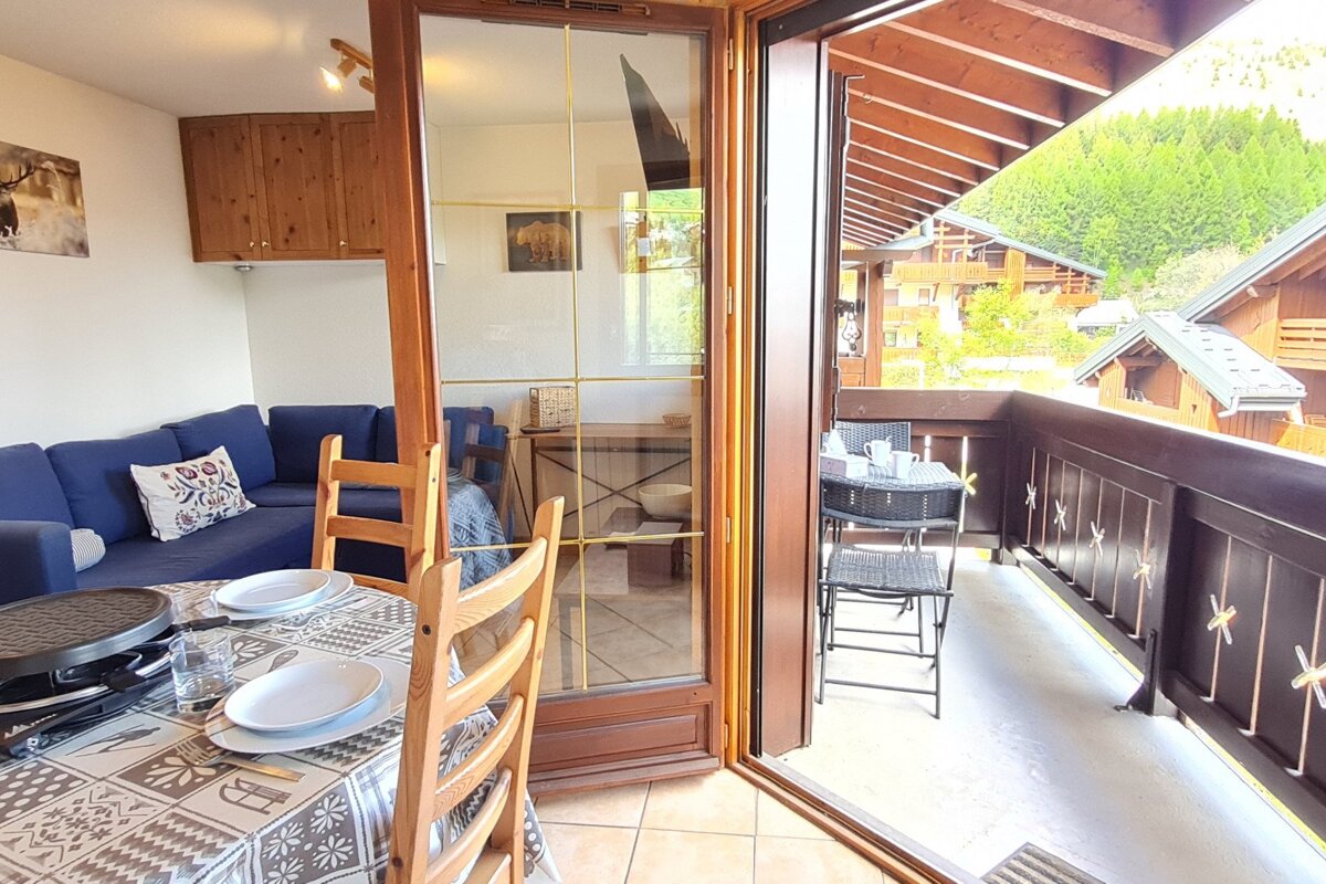 A cozy indoor dining/living area with a blue sofa and raclette grill opens to a balcony with outdoor seating and a view of mountain chalets and trees.