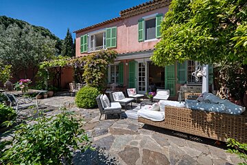 A Mediterranean-style pink house with green shutters and a stone patio featuring outdoor lounge seating amidst lush greenery under a clear blue sky.