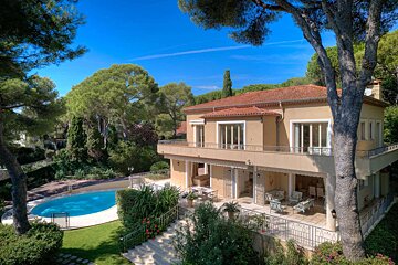 A beautiful Mediterranean villa with a red roof, surrounded by green trees and a clear blue sky, featuring a sparkling swimming pool in the garden.