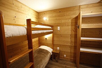 Cozy, wood-paneled room featuring bunk beds, a single bed below, and an open closet with shelves. Warm lighting and rustic charm.