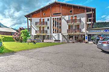 A large, multi-story chalet-style building with white walls, wooden balconies, and stone details, housing a tourism residence and an AXA office, with a lawn and parking.